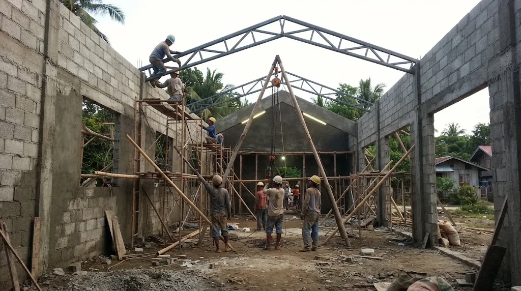 A concrete chapel under construction in a provincial town, with steel roof trusses being lifted into place over the nave