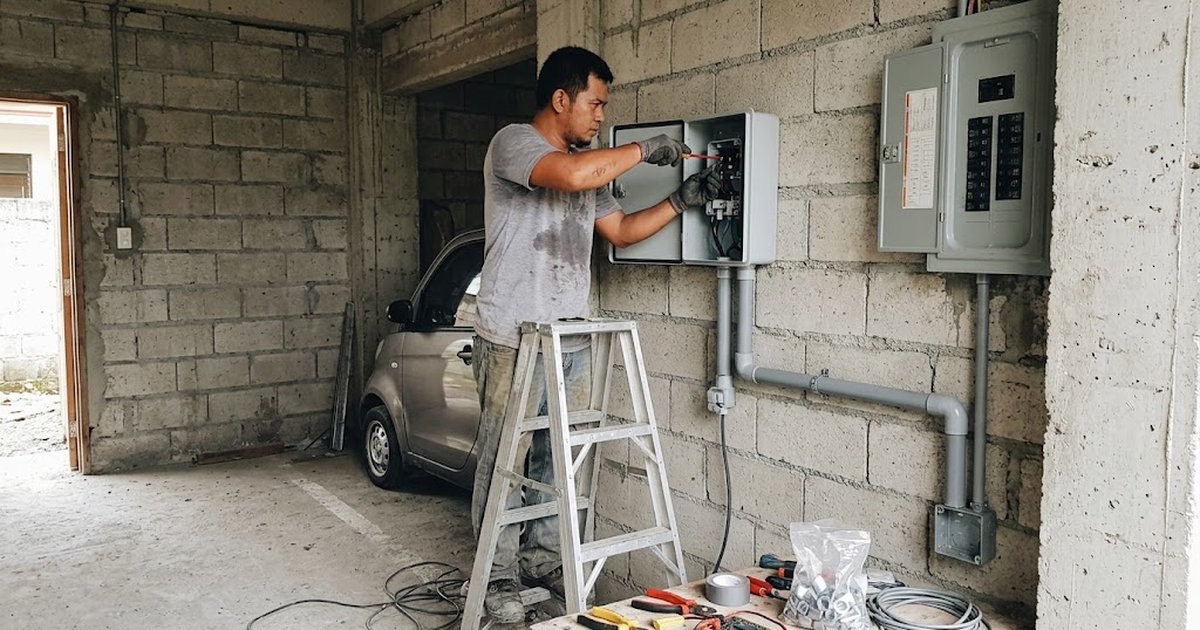 Filipino electrician mounting a wall-box EV charger and running conduit beside a parking slot in a residential garage
