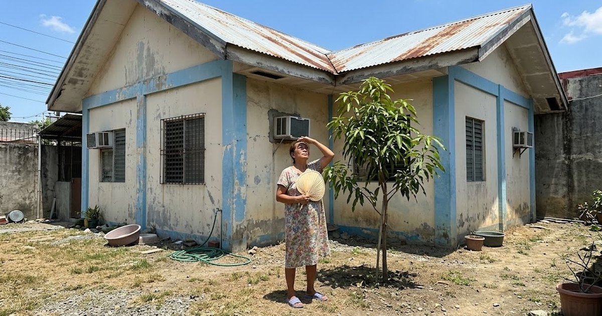 Woman shading her eyes outside a small single-storey house under the midday sun