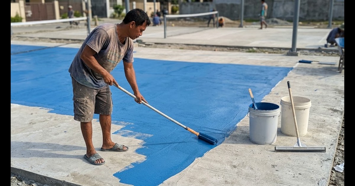 Filipino painter rolling a blue acrylic coat onto a smooth concrete pickleball court slab on a sunny day, paint pails and a squeegee beside him