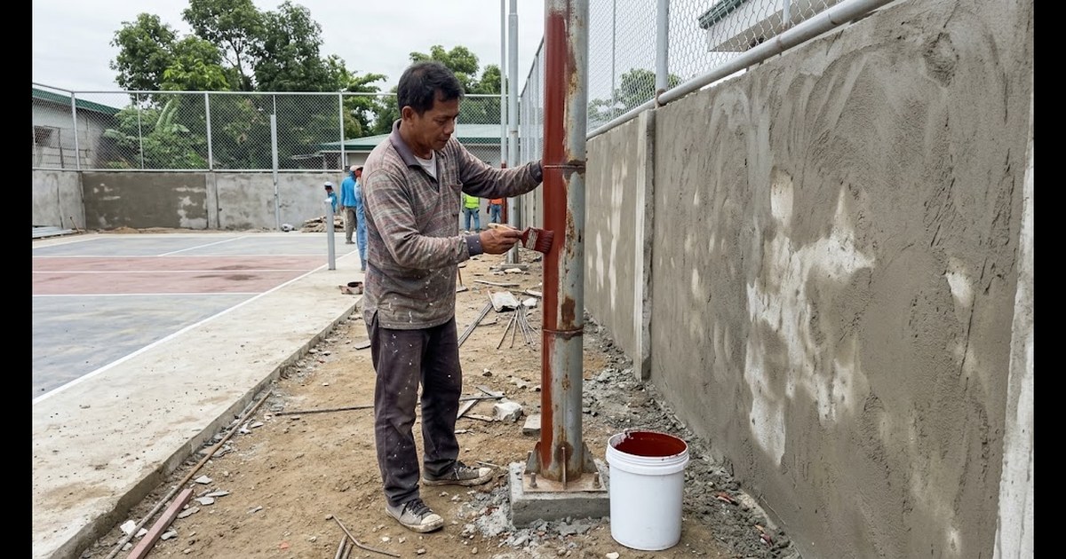 Filipino painter brushing red-oxide primer onto a steel light pole beside a freshly plastered CHB wall at a pickleball court under construction