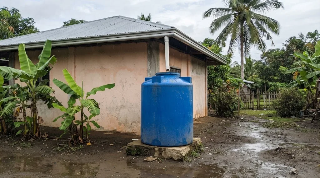 Blue plastic water tank on a concrete base fed by a downspout from the roof gutter of a rural house in the Philippines