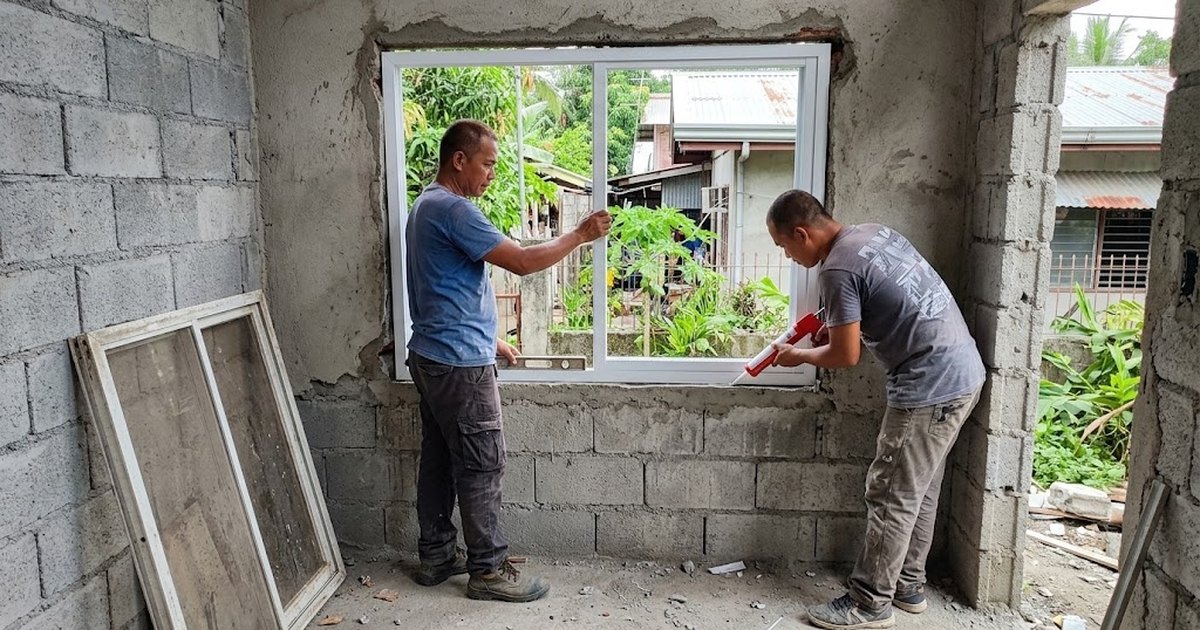 Two workers fitting and sealing a new aluminum sliding window in a hollow block wall